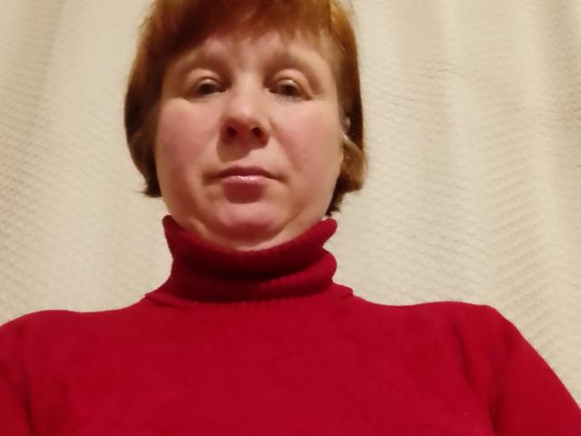 JackFine hat kurze, braune Haare, einen hellen Hautton und trägt einen roten Rollkragenpullover. Sie hat ein leichtes Lächeln und ein leicht faltiges Gesicht. Ihre Brüste sind nicht sichtbar..