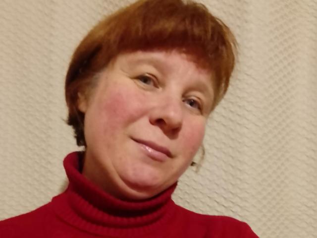 JackFine tem cabelo ruivo curto, um rosto levemente avermelhado e está vestindo um cachecol vermelho. Ela está sorrindo suavemente, com uma leve inclinação na cabeça..