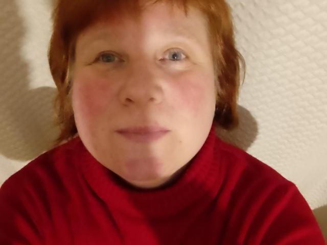 JackFine hat rotes Haar, blaue Augen und trägt einen roten Rollkragenpullover. Ihr Gesicht ist leicht unscharf, mit einem zarten, rosa Schimmer und einem neutralen Ausdruck..
