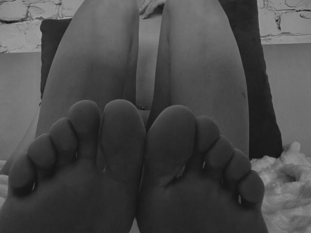HollyHope's bare feet with toes pointed upward, her soft pussy partially visible between her thighs, lying on a bed with a dark pillow behind her. Black-and-white image.