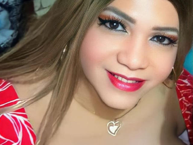 Kelly, com longos cabelos castanhos, vestindo um vestido floral vermelho e branco, brincos de argola dourados, um colar de coração e batom rosa vibrante, sorrindo para a câmera com delineador marcante..