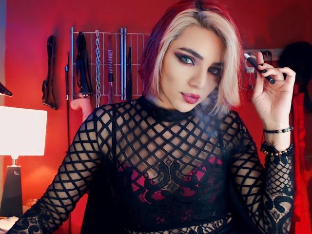 MisssDarkLady, avec ses cheveux blonds, son haut en résille noire laissant entrevoir ses seins, son rouge à lèvres rouge et son maquillage des yeux fumé, tenant un rouge à lèvres noir, dans une pièce rouge avec du matériel BDSM..