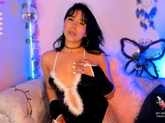 ElizabethMacy with black hair, wearing a black velvet bodysuit with white fur trim, fishnet stockings, black choker, and fingerless gloves, holding her tits, sitting on a white couch.