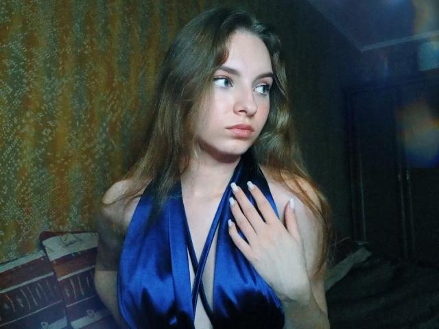 MissDi com cabelos castanhos longos, blusa de alça de cetim azul, mão no peito, expressão emburrada, pele clara, maquiagem discreta, olhando para o lado, em um quarto com pouca luz..