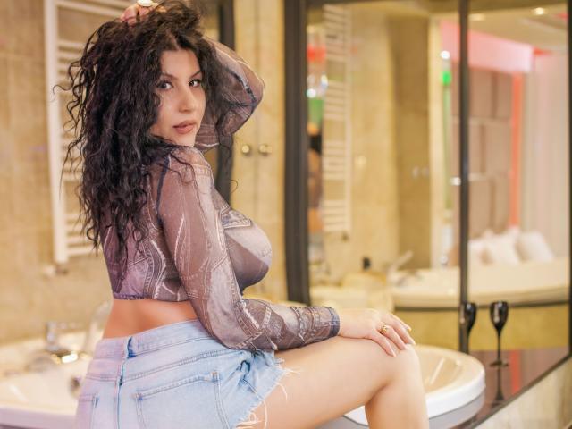 AddaCurvyHot mit lockigem, schwarzem Haar, die ein durchsichtiges, lila Oberteil und zerrissene, blaue Jeansshorts trägt, sitzt auf einem Badezimmerwaschbecken, blickt über ihre Schulter und zeigt ihre Brüste und ihren Hintern..