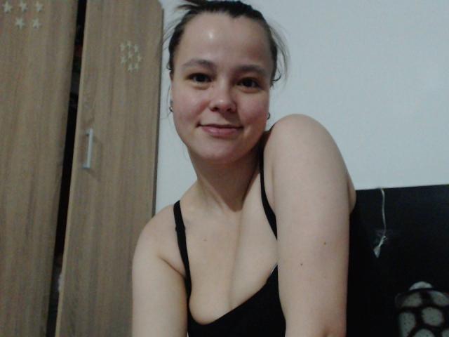 Una mujer llamada HotJoyLover, de piel clara y cabello oscuro, vestida con una camiseta negra, sonríe, tiene un brazo levantado mostrando su hombro descubierto y sus pequeños senos..