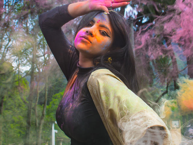 CoriPaige with colorful face paint, black top, and gold sleeves, arm raised, purple-pink tree in background, green foliage, and pink-orange light on her face.