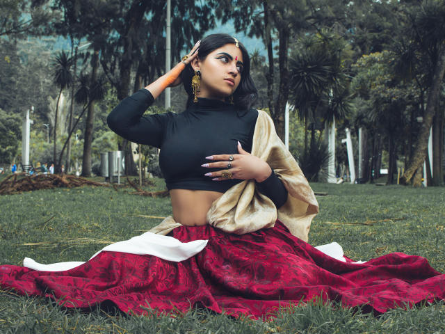 CoriPaige has big tits, wearing a black crop top and red patterned skirt with a beige shawl, sitting on grass, hand on head, other hand on tits, forest background.