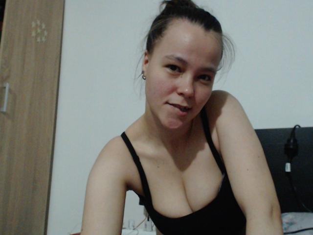 HotJoyLover with brown hair in a bun, black tank top, showing medium-sized tits, slightly leaning forward, smiling, nose ring, and small ear gauges.