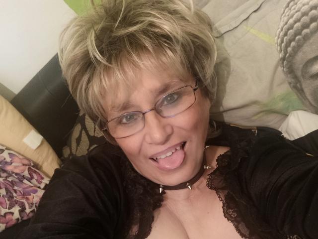 Samantha Mature hat kurze, blonde Haare, eine Brille und eine schwarze Halskette. Sie steckt ihre Zunge heraus, und ihre Brüste sind in einem Spitzenoberteil sichtbar. Sie sieht verspielt aus..