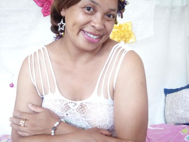 Rahel69 with brown skin, short brown hair, and star earrings, smiling in white lace top, arms crossed, pink lipstick, ring on right hand, bracelet on left wrist.