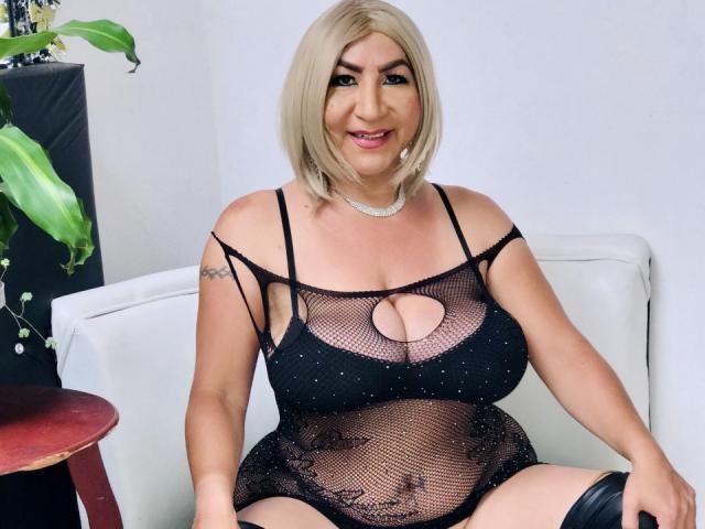 TsMonterCock with blonde bob, wearing a black, see-through mesh dress, showing her large boobs and belly, sitting on a white chair, smiling, with a tattoo on her arm.