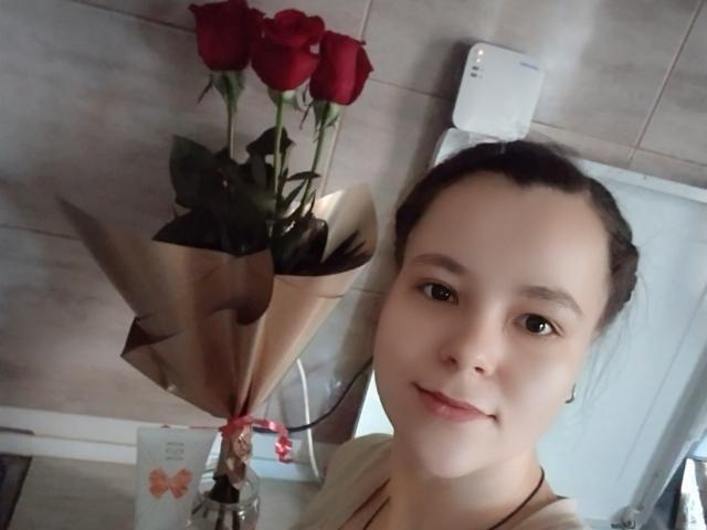 HotJoyLover com pele clara, cabelo castanho preso em um coque, sorrindo, segurando um buquê de rosas vermelhas com folhas verdes e papel marrom, em um banheiro..