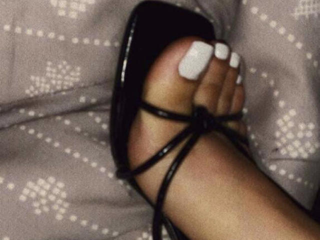 StormyKiss' Fuß mit weißem Nagellack auf dem großen Zeh, trägt einen schwarzen, hohen Sandalettenabsatz und ruht auf einer gemusterten Oberfläche..