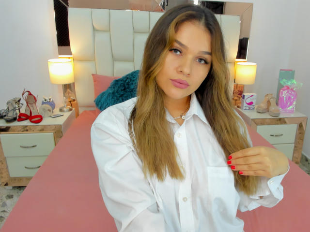 ValerieStone with long brown hair, pink lips, wearing a white shirt, red nails, standing in a bedroom, holding shirt, bed with pink blanket and white headboard in background.