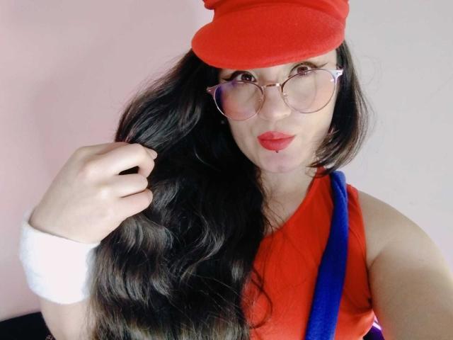 MillaDrew, con cabello largo y negro, usando un gorro rojo, gafas, una camiseta sin mangas roja, una pulsera blanca y una correa azul, haciendo pucheros con lápiz labial rojo, sosteniendo su cabello..