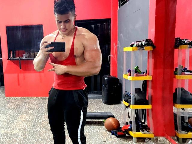 AngelLin, com bíceps enormes, brilhantes e musculosos, vestindo uma regata vermelha e calças de moletom pretas, tirando uma selfie em uma academia, segurando um celular com uma expressão séria..