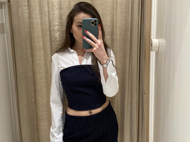 Dionisia with long brown hair, wearing a white shirt and dark blue pinstriped tube top and skirt, holding a green phone, showing red nails, and a belly button piercing.