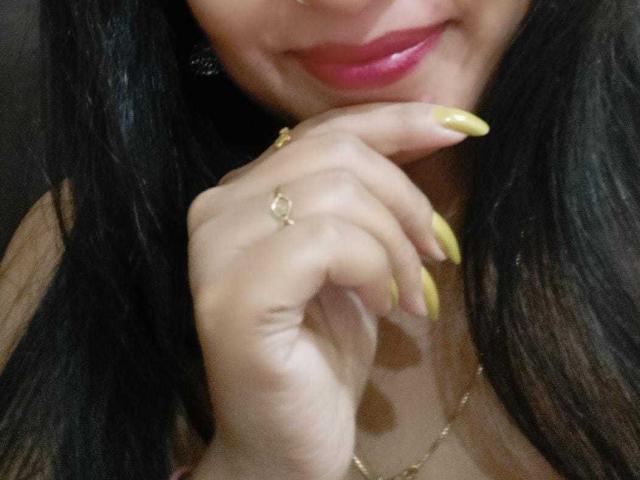 La main d'ElizaNdKeo69, avec ses ongles jaunes, sa bague et son collier, touche ses lèvres légèrement entrouvertes, révélant un sourire subtil. Elle a de longs cheveux noirs et un anneau au nez..
