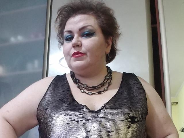 KaryQueen with large tits, pale skin, curly brown hair, blue eyeshadow, red lipstick, black sequined tank top, multiple chain necklaces, standing indoors, looking at camera.