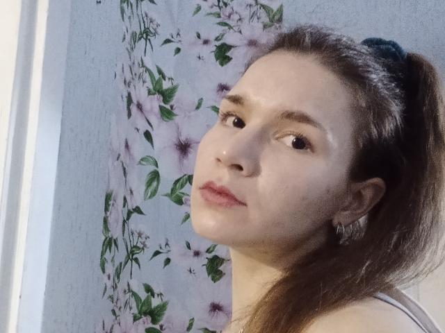VeraKyk has shiny skin, brown eyes, and dark brown hair in a ponytail, wearing a white top, with floral earrings and a subtle smile.
