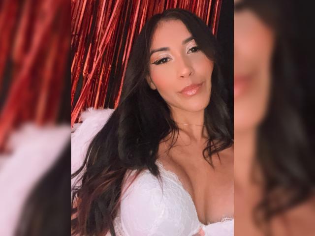AnyaJay with long black hair, white lace dress, and large boobs, smiling softly at the camera, red ribbon-like background blurred on the left and right.