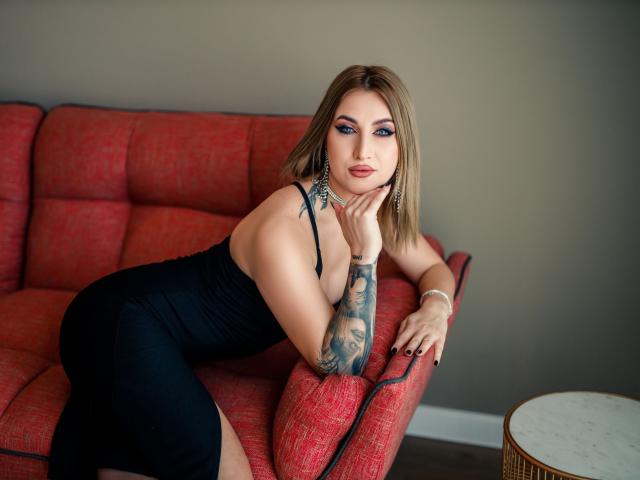 LexieSpark, met blauwe oogmake-up, een zwarte jurk en tatoeages, leunt tegen een rode bank, met haar hand op haar wang, en laat haar borsten en billen zien, terwijl ze naar de camera kijkt, met haar arm op de bank..