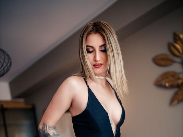 LexieSpark, met blond haar, draagt een diepe zwarte jurk, waarbij haar borsten zichtbaar zijn door de lage halslijn, waardoor haar choker en de tatoeage op haar rechterarm te zien zijn. Ze kijkt naar beneden..