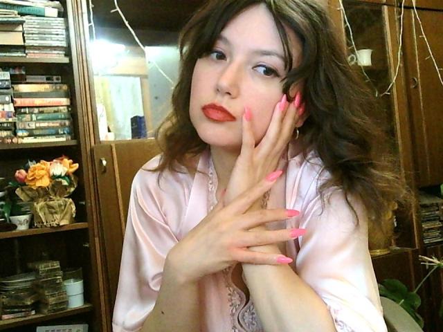 CarriePeachy, met roze nagels, draagt een roze satijnen shirt, heeft golvend bruin haar, rode lippenstift en een bedachtzame uitdrukking, waarbij ze haar wang aanraakt met haar rechterhand..