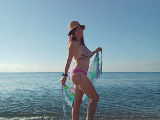 Cutematurelatin with long brown hair, tan skin, wearing a patterned bikini, pink thong, straw hat, and holding a colorful scarf, standing in shallow ocean water, smiling.