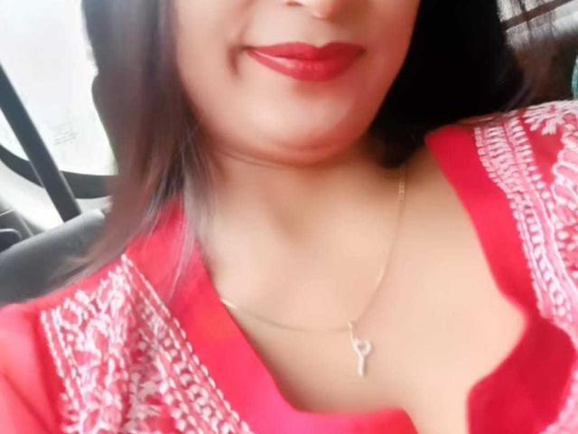 ElizaNdKeo69 con rossetto rosso, sorridente, indossa una maglia rossa con motivi bianchi e una collana con una chiave dorata, con un leggero spacco che mostra il décolleté. I suoi capelli castani sono parzialmente visibili..