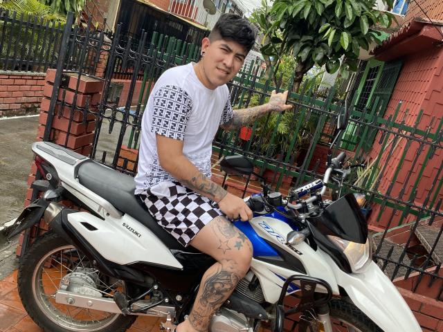 CrazyTatto met tatoeages, een zwart-wit geruit shirt, zwarte en witte geruite korte broek, zittend op een witte en blauwe Kawasaki motorfiets, en een duimpje omhoog..