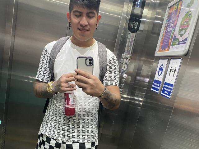 CrazyTatto, avec ses cheveux noirs courts, des tatouages sur les deux bras, portant une chemise blanche et noire à motifs, un pantalon à carreaux, un sac à dos et une montre en or, prend un selfie dans un ascenseur métallique..