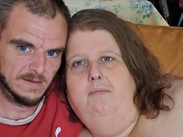 Het mollige gezicht van StephanieLeo69, met bleke huid en blauwe ogen, naast een man met een baard en een rood shirt, beiden met kort bruin haar, liggend op een geel kussen..