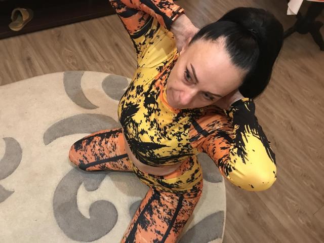 RoxolanaXDrim, avec ses cheveux noirs attachés en queue de cheval, portant une tenue jaune et orange avec un motif éclaboussé, est agenouillée sur un tapis, les mains derrière la tête, et regarde la caméra..