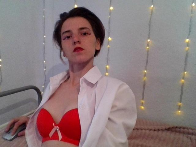 RoseJill met kort donker haar, een rode bh die haar borsten laat zien, een wit shirt dat open staat, zittend op een bed, met rode lippenstift, kerstverlichting op de achtergrond, kijkend naar de camera..