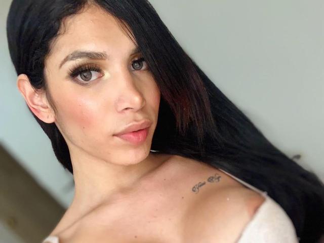ExoticHorny hat langes, schwarzes Haar, braune Augen und ein Tattoo an ihrer rechten Schulter. Sie trägt ein weißes Oberteil, das ihre kleinen Brüste und ihre rosa Lippen zeigt..