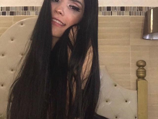 ExoticHorny met lang zwart haar, lachend, haar borsten showend en er schattig uitzien, zittend op een beige bed met een gouden bedstijl..