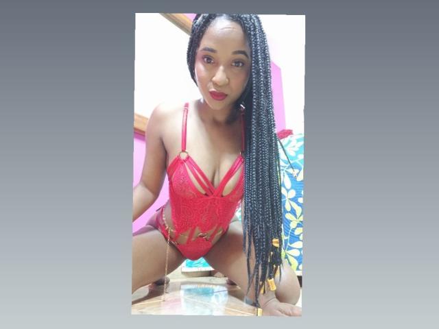 MelanyScoth with long black braids, wearing a red lace lingerie top with cutouts, squatting, showing her small tits and bare pussy, looking at the camera with red lipstick.
