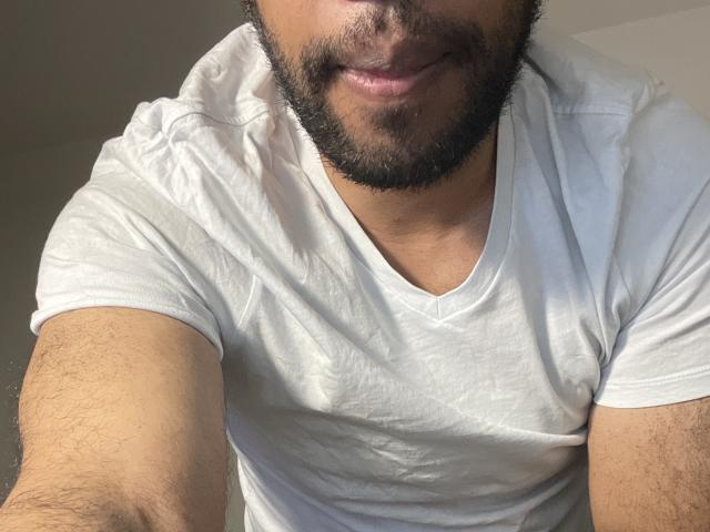 Olhivia, avec une barbe, un t-shirt blanc et des poils de poitrine légèrement visibles, sourit à l'appareil photo. Sa peau est bronzée et sa barbe est foncée et bien taillée..