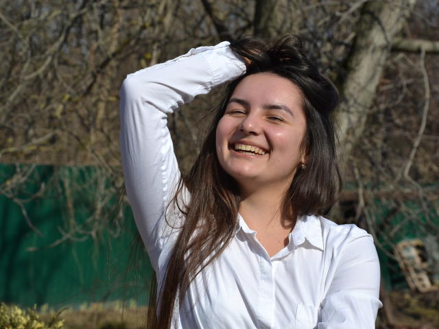 Jasmine Asha mit langen, braunen Haaren, lächelnd, trägt ein weißes Hemd mit Knopfleiste, hebt ihren rechten Arm, zeigt ihr Gesäß, steht draußen mit einem grünen Zaun im Hintergrund..