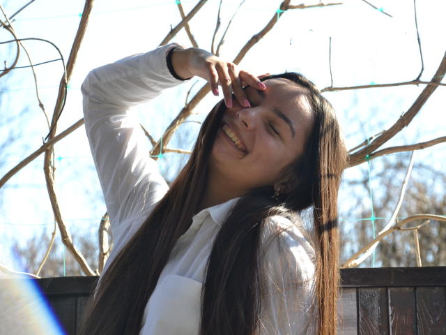 JasmineAsha with long brown hair, smiling, eyes closed, white shirt, hand touching face, sunlight, wooden fence, leafless branches, happy expression, relaxed, natural light, casual, daytime.