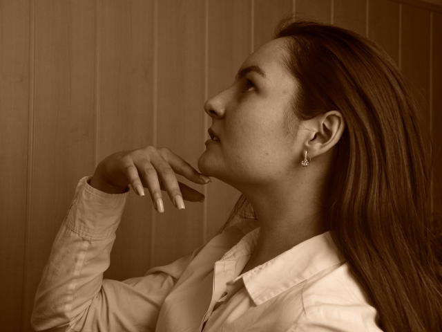 Sepia-toned photo of JasmineAsha with long brown hair, wearing a white shirt, looking up, hand near chin, earrings visible, serious expression, slight smile, soft lighting.
