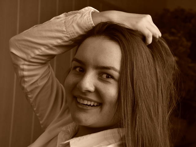 Sepia-toned photo of JasmineAsha with long brown hair, smiling, wearing a white shirt, hand in hair, showing cute teeth, and slightly freckled face. Boobs not visible.
