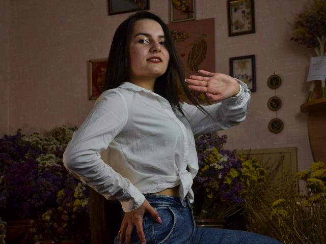 JasmineAsha, with dark hair, wearing a white button-up shirt tied at the front and blue jeans, is waving her right hand, showing off her small tits.