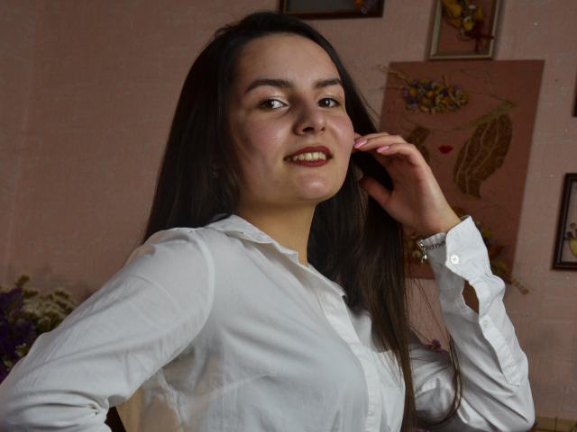 JasmineAsha with long dark hair, white button-up shirt, medium tits, slight smile, hand near face, soft pink skin, brown eyes, standing indoors, framed art in background.