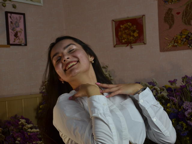 JasmineAsha, avec ses longs cheveux noirs, souriant les yeux fermés, les mains près de son cou, portant une chemise blanche, dans une pièce remplie de fleurs, montrant son joli visage et sa poitrine..
