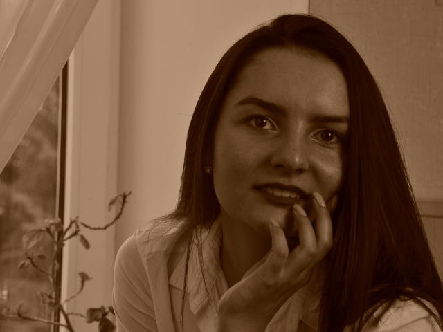 Fotografia in bianco e nero di JasmineAsha con lunghi capelli scuri, che si morde un dito, indossa una camicia bianca, guarda leggermente di lato con un sorriso appena accennato, mostrando i suoi occhi seducenti..