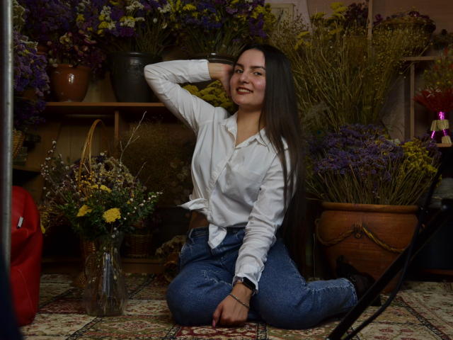 JasmineAsha with long dark hair, white shirt, and blue jeans, kneeling, smiling, hand in hair, surrounded by flowers, wearing a bracelet, and a watch, in a floral-filled room.