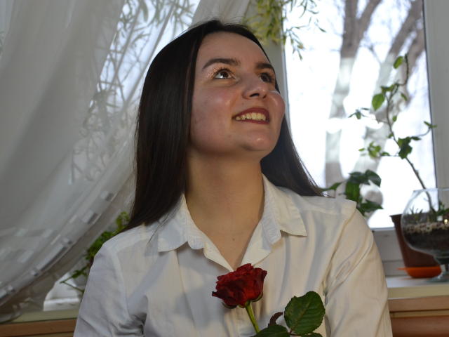 Jasmine Asha mit langem, schwarzem Haar, lächelnd, trägt ein weißes Hemd, hält eine rote Rose und steht in der Nähe eines Fensters mit weißen Vorhängen und grünen Pflanzen..